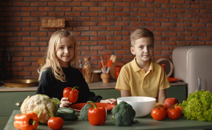 Positive little girl and boy sitting at table with various vegetables and herbs while preparing vegetarian meal in loft kitchen with brick wall